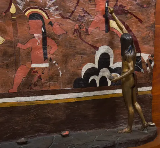 Bronze statue of a naked man with long black hair, wearing sandals, raising one arm toward an earthy-toned mural on a brown wall.