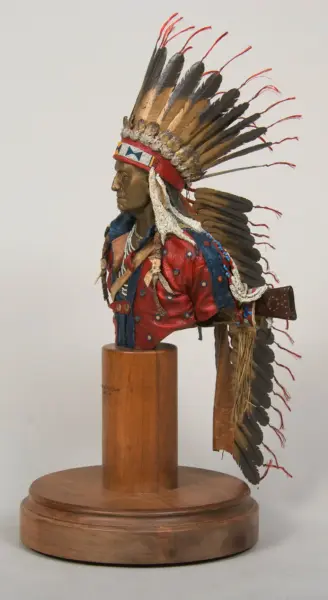 Bust of a Native American man wearing a tall feathered war bonnet, red-and-blue beaded clothing, and layered jewelry, mounted on a wooden pedestal