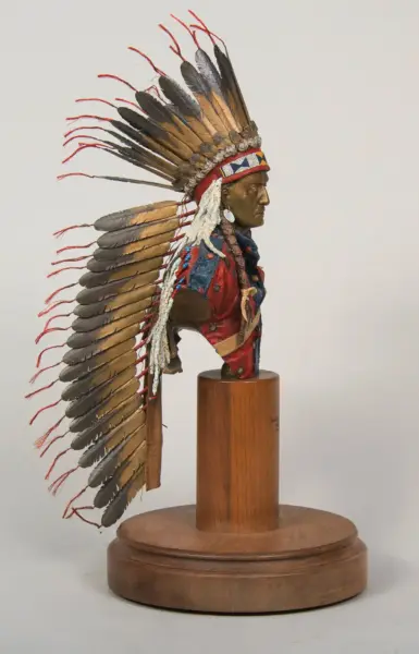 Bust of a Native American man wearing a ceremonial feather headdress on a wooden pedestal display.