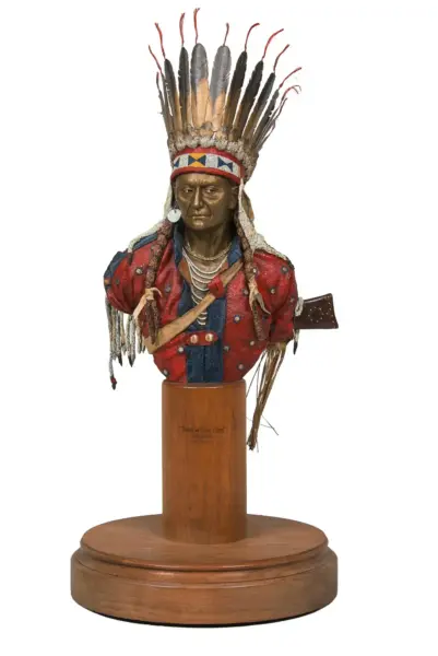 Bust of a Native American chief wearing a tall feather headdress and beadwork, displayed on a wooden pedestal
