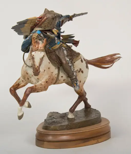 Elaborate sculpture of a warrior riding a spotted horse, wearing decorative attire and a winged headdress, aiming a rifle on a wooden base.