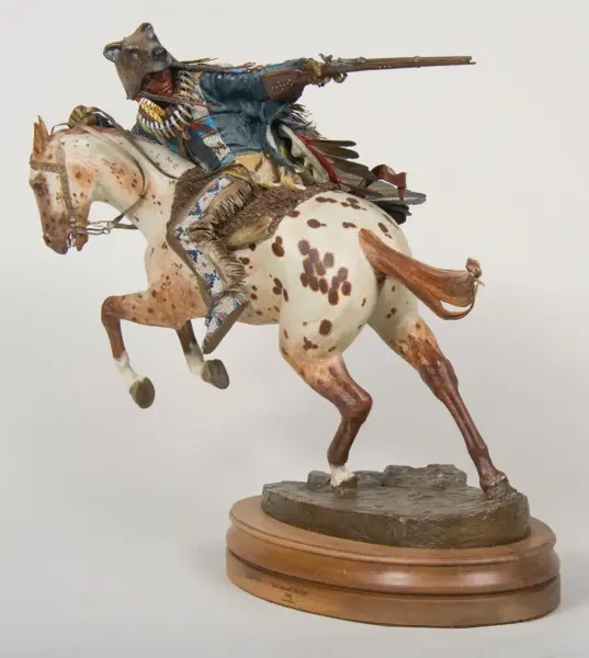 Detailed sculpture of a Native American rider on a rearing white-spotted horse, aiming a long rifle, wearing fringed blue clothing and beadwork on a wooden base