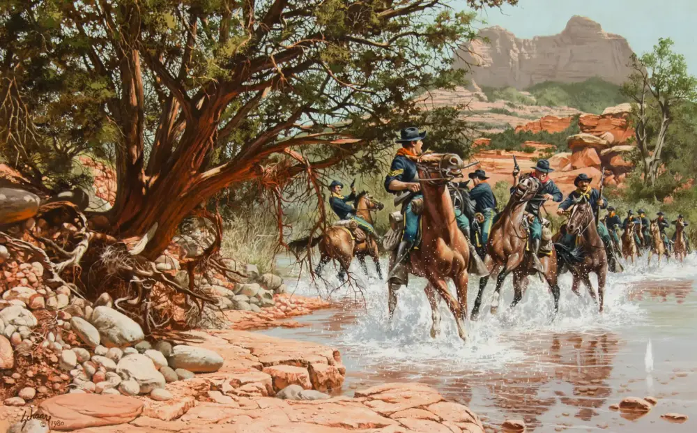 Cavalry riders in blue uniforms cross a shallow river amid a desert canyon landscape with red rock formations and a large tree in the foreground.