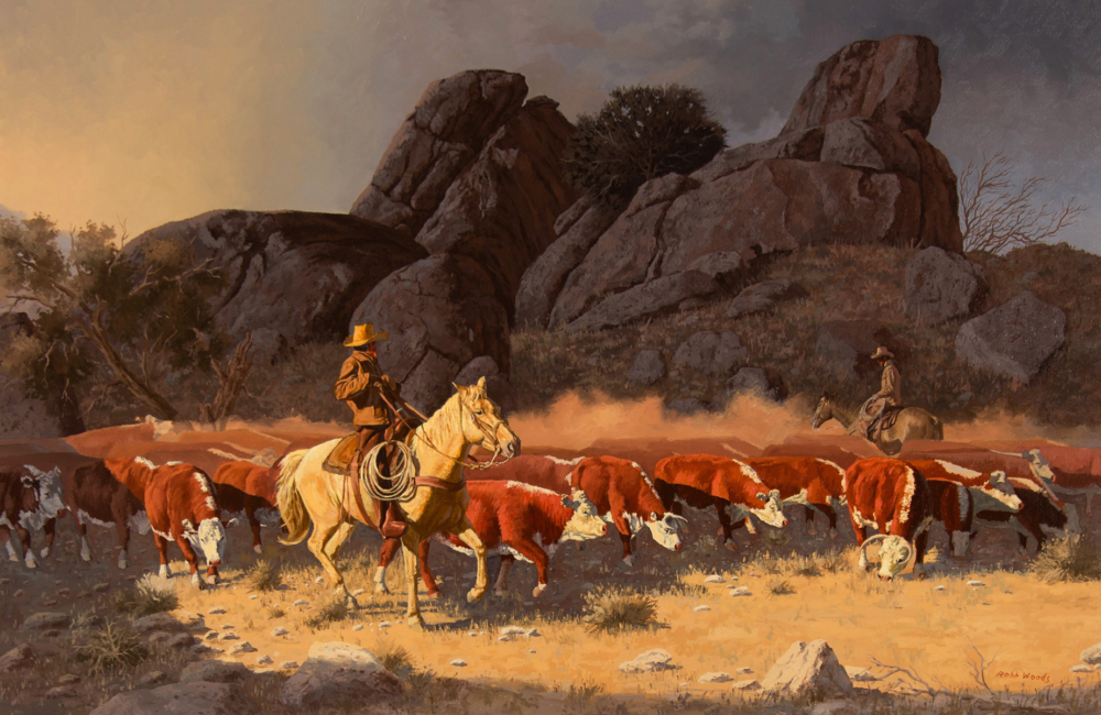 Two cowboys on horseback lead a long herd of red-and-white cattle across a dry, rocky plain with large desert boulders in the background.