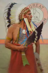 Native American man in a tall feather headdress and red ceremonial robe, holding a large feathered staff against a pale mural backdrop.