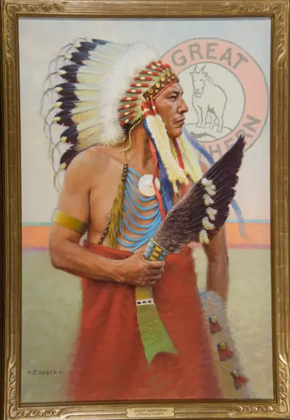 Portrait of a Native American man in a colorful feather headdress holding a feathered staff, with a Great Northern logo in the background.