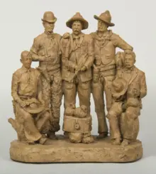 Clay sculpture of six men in old-time western outfits standing together on a base, with hats and rugged clothing.