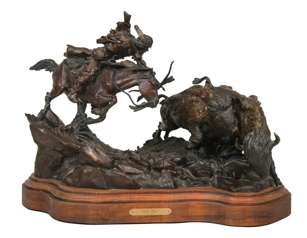 Bronze sculpture of a rider on a rearing horse chasing a buffalo across jagged rocks, mounted on a wooden base with a plaque