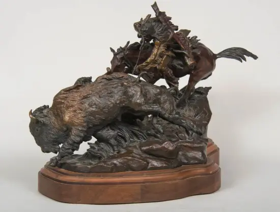 Bronze sculpture of a charging bison with a rider clinging to its back on a rocky pedestal.