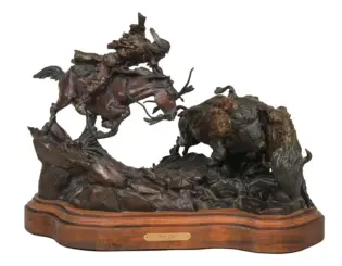 Bronze sculpture of a rider on a rearing horse chasing a buffalo across jagged rocks, mounted on a wooden base with a plaque