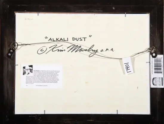 Back of a framed artwork showing the handwritten title 'ALKALI DUST', with the artist signature 'Kim Mackey' and hanging wires secured to the frame; includes small museum labels and a portrait card attached to the lower left.