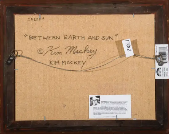 Back of a framed painting shows handwritten title 'Between Earth and Sun' by Kim Mackey with a hanging wire and label tags nearby.