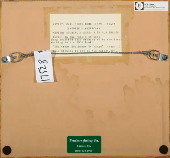 Back of a framed artwork showing a typed label about artist Carl Oscar Borg and the etching 'In the Pueblo of Taos'; a metal hanging wire with hooks runs across, with a white numbered tag hanging from it, and a green Freelance Gilding Inc. sticker at the bottom.
