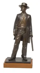 Bronze statue of a cowboy in a wide-brimmed hat and long coat, holding a pistol, mounted on a wooden base.