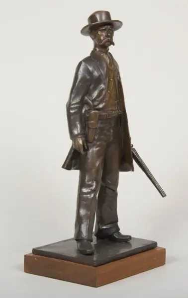 Bronze statue of a man in a wide-brimmed hat and long coat, with a sword by his side, standing on a wooden base.
