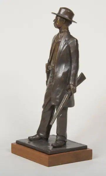 Bronze statue of a man in a hat and suit, standing with a rifle by his side on a wooden base.