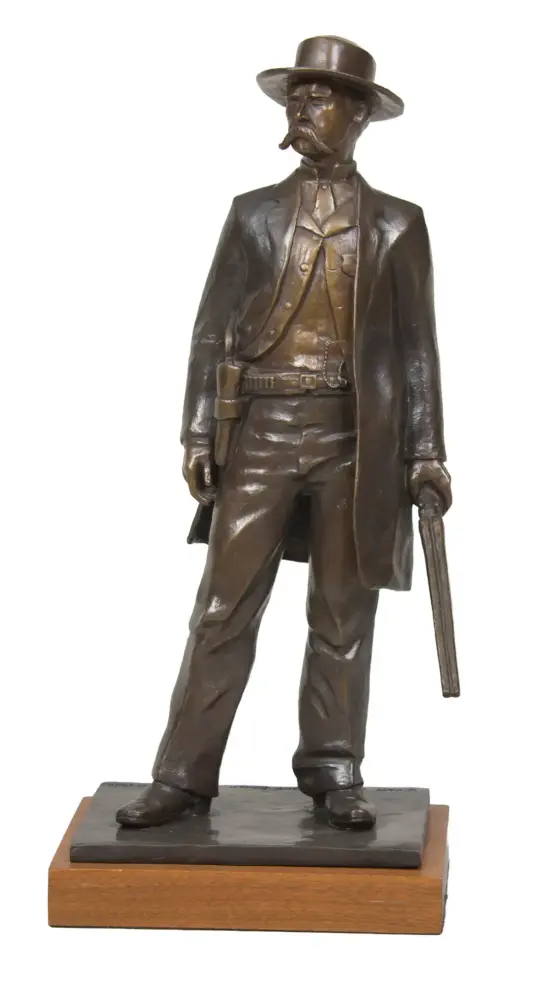 Bronze statue of a cowboy in a wide-brimmed hat and long coat, holding a pistol, mounted on a wooden base.