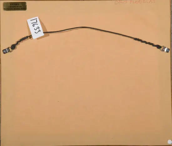 Back of a light brown mounting board with a twisted black cord and metal clasps at each end, plus a white tag labeled '7633' pinned near the left.