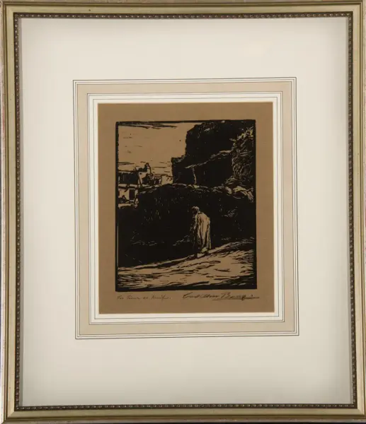 Framed vintage-style print showing a coastal cliff scene with a person standing near rocks on a shoreline, mounted on layered tan and white mats in an ornate frame.