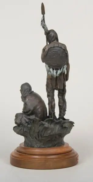 Bronze sculpture of a standing Native American man raising a spear above his head, with a shield on his back and feathered belt, beside a seated figure, set on a wooden pedestal.