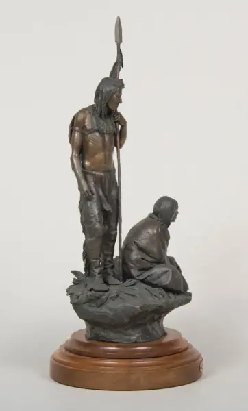 Bronze sculpture of a standing Indigenous man with a tall spear, beside a seated figure, on a rocky pedestal with a wooden base.