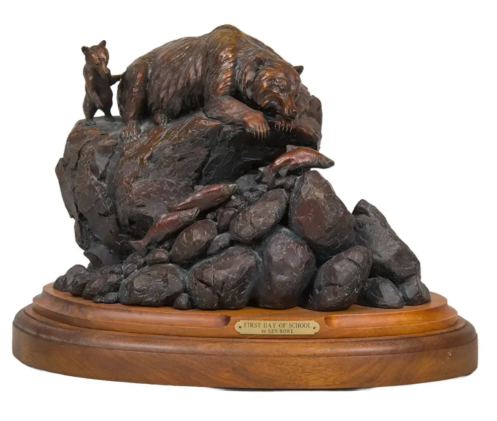 Bronze-toned sculpture of a mother bear and cub on a rocky outcrop atop a wooden pedestal with a plaque reading 'First Day of School'.