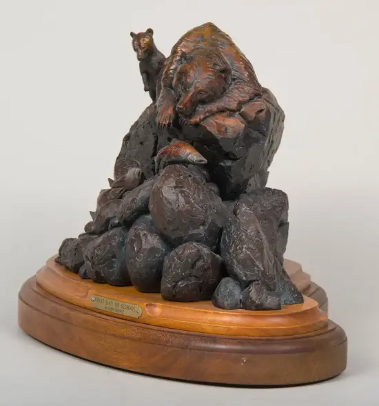 Bronze-toned sculpture of a bear and cubs resting on a jagged rock formation atop a wooden pedestal, with a plaque reading 'First Day of School' by Ken Rowe at the base.