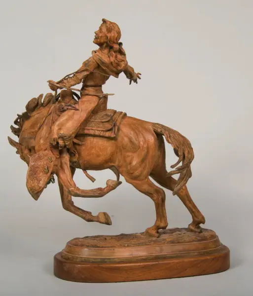 Bronze-colored sculpture of a cowboy riding a bucking horse on a round wooden pedestal, mid-action pose.