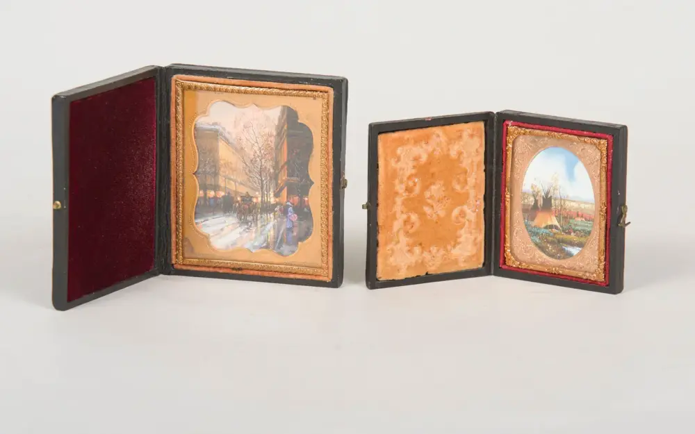 Open antique black folding photo case with burgundy velvet interiors; left panel displays a winter street scene in a decorative gold frame, middle panel blank, right panel shows a pastoral landscape photo in a gold-toned frame.