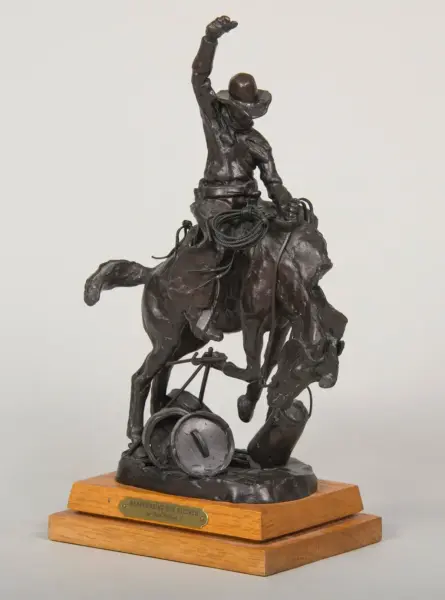 Dark metal sculpture of a cowboy on a rearing horse, one arm raised, with ropes and gear, on a wooden base.