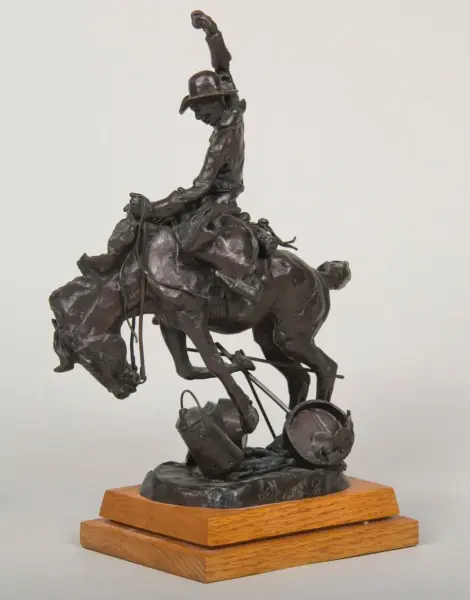 Bronze sculpture of a cowboy riding a bucking horse, hat on, raised arm, on a wooden pedestal base.