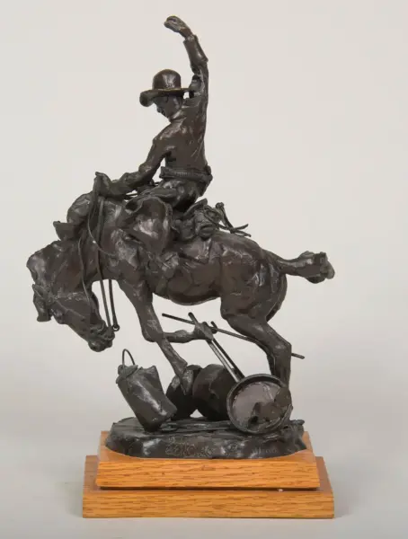 Bronze sculpture of a cowboy riding a rearing horse, hat tipped, one arm raised, mounted on a wooden base