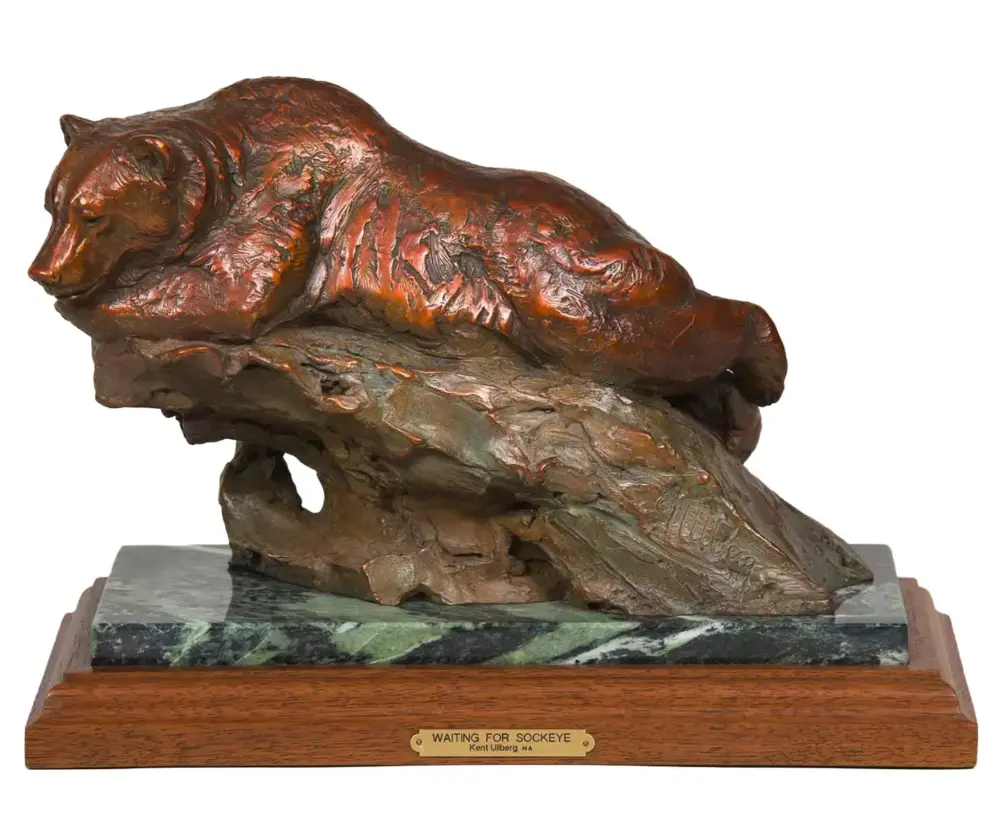 Bronze bear statue lying on a rock, mounted on a wooden base with a plaque reading 'Waiting for Sockeye'.