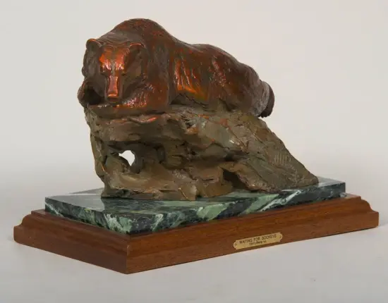 Bronze bear sculpture resting on a rock pedestal, mounted on a green marble base with a wooden platform and a small plaque reading 'Waiting for Sockeye'.