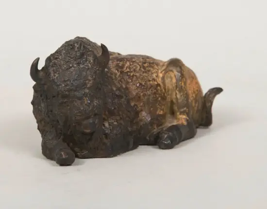 Bronze sculpture of a lying-down bison with curved horns and a rugged, textured surface.
