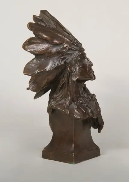 Bronze bust of a person wearing a layered feathered headdress, facing right, on a square pedestal.