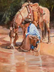 Man in a light blue shirt and black hat kneels by a horse at a muddy watering hole.