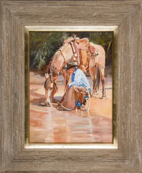 A person kneels beside two saddled horses drinking from a shallow pool, in a painted outdoor scene framed in a rustic wooden frame.