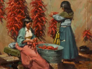 Two women work with red chili peppers: one seated peeling peppers into a tub, the other standing and tying peppers on a string as peppers hang nearby.