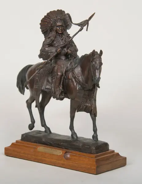 Bronze statue of a Native American warrior on horseback, wearing a feathered headdress, on a wooden base with a plaque.