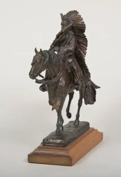 Bronze statue of a Native American rider on a horse, wearing a feathered headdress, mounted on a wooden base.
