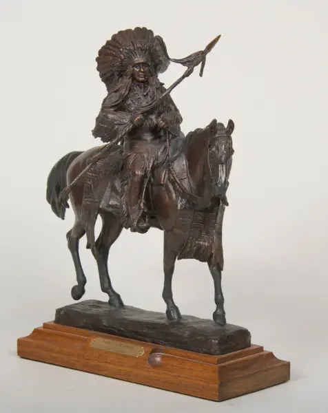 Bronze statue of a Native American warrior on horseback, wearing a feather headdress and holding a spear, mounted on a wooden base.