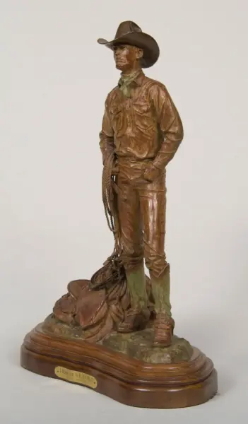 Bronze statue of a cowboy in a hat, with a lasso at his side, standing on a wooden base.