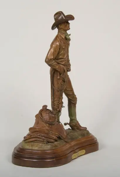 Bronze statue of a cowboy in a wide hat, standing with hands on hips, rope coiled at his side on a rocky base with a saddle nearby.