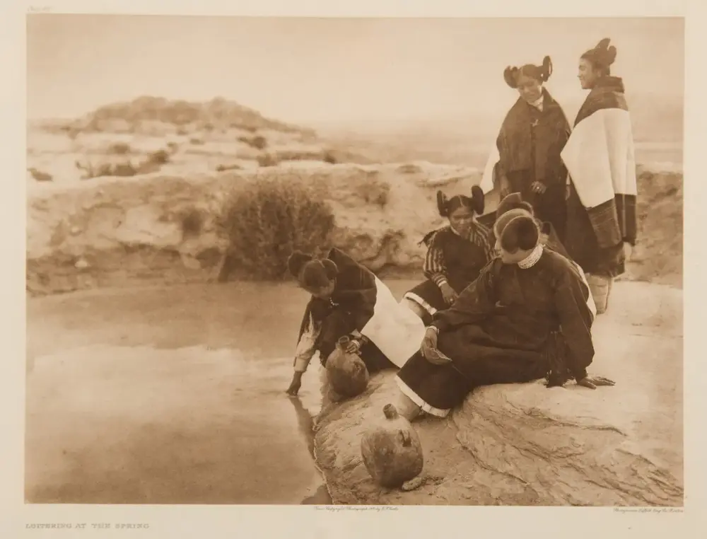 Indigenous people in traditional dress at a rocky spring; a woman sits with a clay water jug while children gather nearby, and others stand in the background.