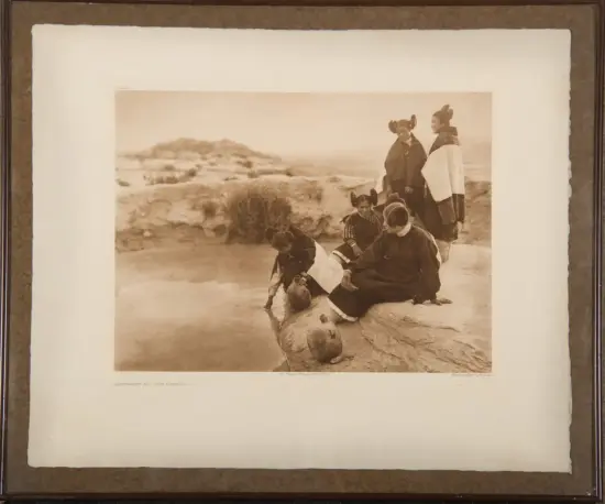 Sepia-toned photo of Indigenous women and children at a rocky spring, dipping pots into the water while two figures in cloaks stand nearby.