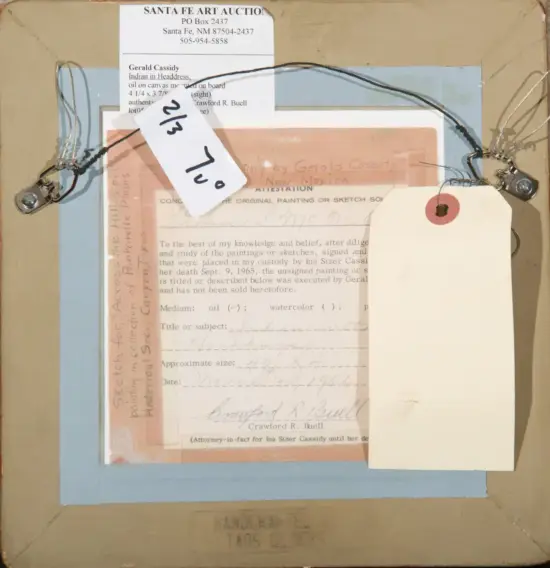 Back view of a framed artwork with layers of labels and tags: a Santa Fe Art Auction label at top, a pale pink document beneath a light blue mat, and a blank white tag attached by string to the frame; metal fasteners visible on the sides.