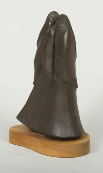 Dark sculpture of two figures embracing, cloaked in flowing robes, mounted on a wooden base.