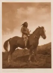 Sepia-toned photograph of an old-time Native American warrior bare-chested, riding a horse across a grassy hillside, holding a spear.