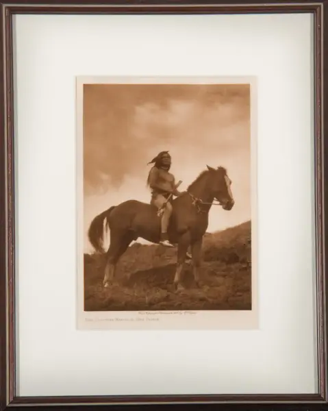 Sepia-toned photograph of a Native American man riding a horse on a hillside, holding a rifle, framed as a vintage print in a dark wooden frame.